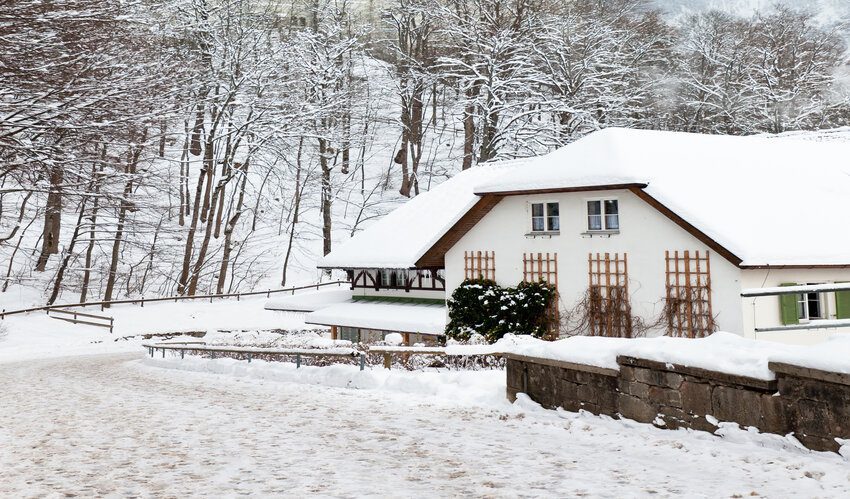 heavy snow over the roof on a white exterior house with trees on the left side of the picture